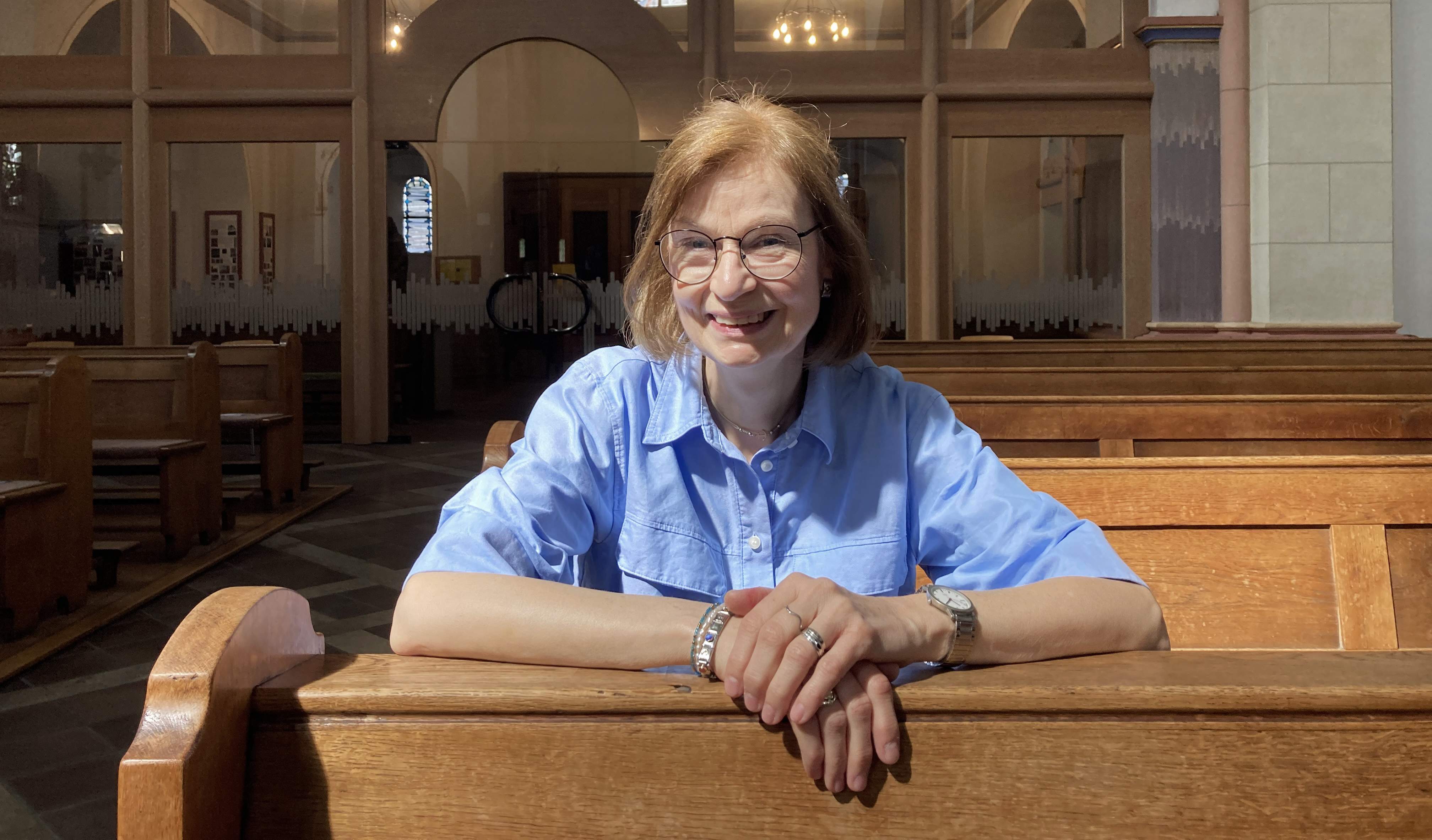 Eine Frau sitzt in einer Kirchenbank und lächelt in die Kamera.