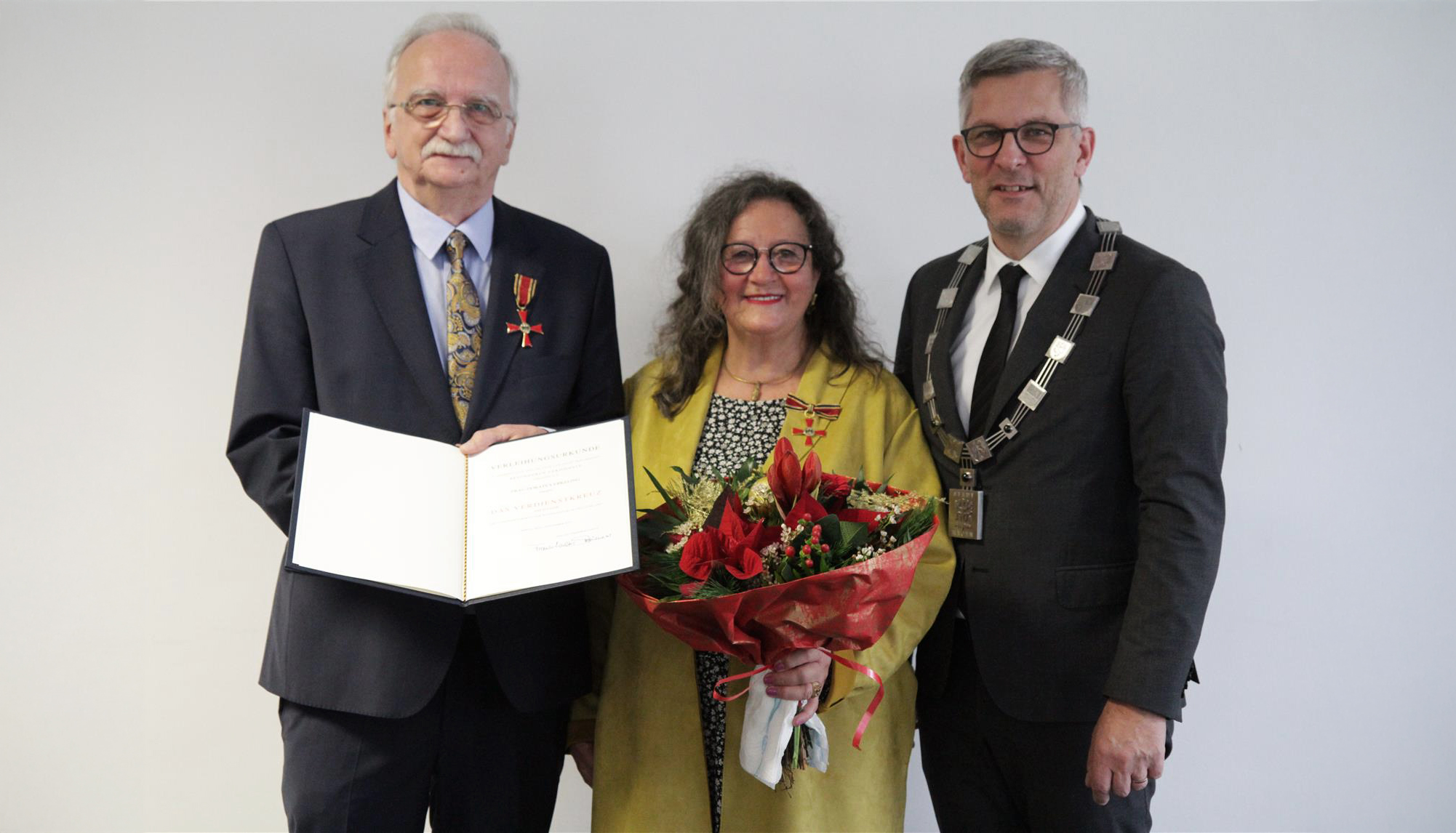 Das Bild zeigt zwei Männer und eine Frau. Der ältere Herr hält eine Urkunde ins Foto. Die Frau, Doratea Erkeling, hält einen Blumenstrauß.