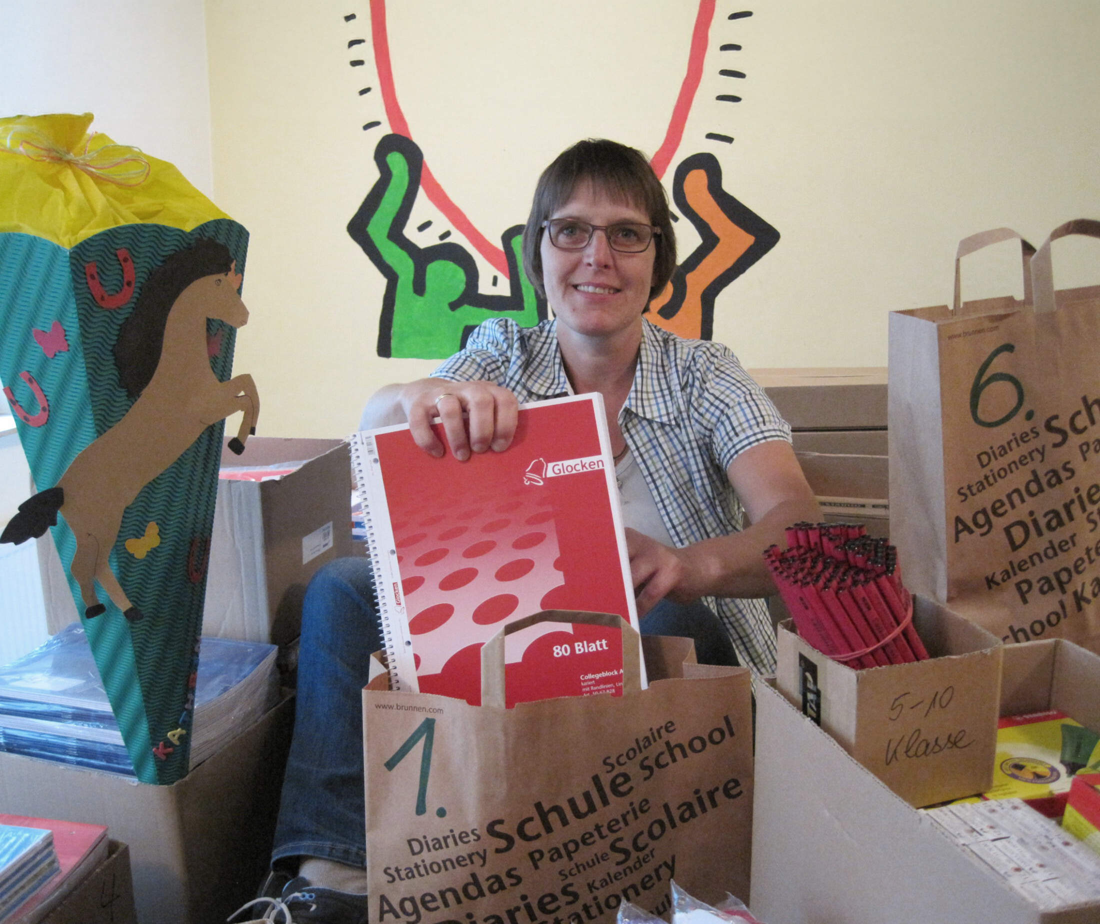 Das Bild zeigt eine Frau, die inmitten von gepackten Tüten mit Schulmaterialien sitzt.