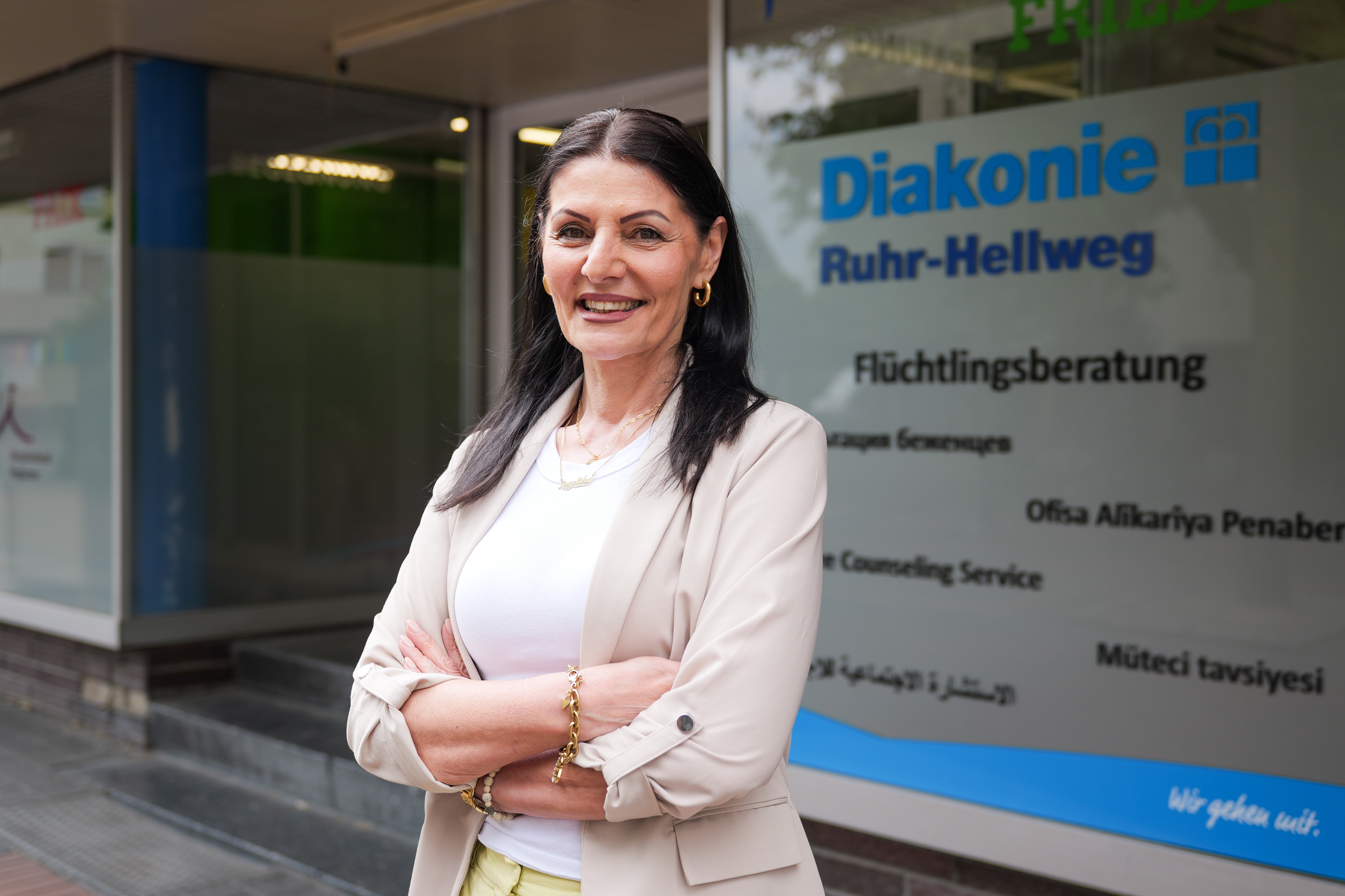 Eine Frau steht vor einem Büro der Diakonie. Sie hat schwarze Haare und trägt einen beigen Blazer über einem weißen Hemd.