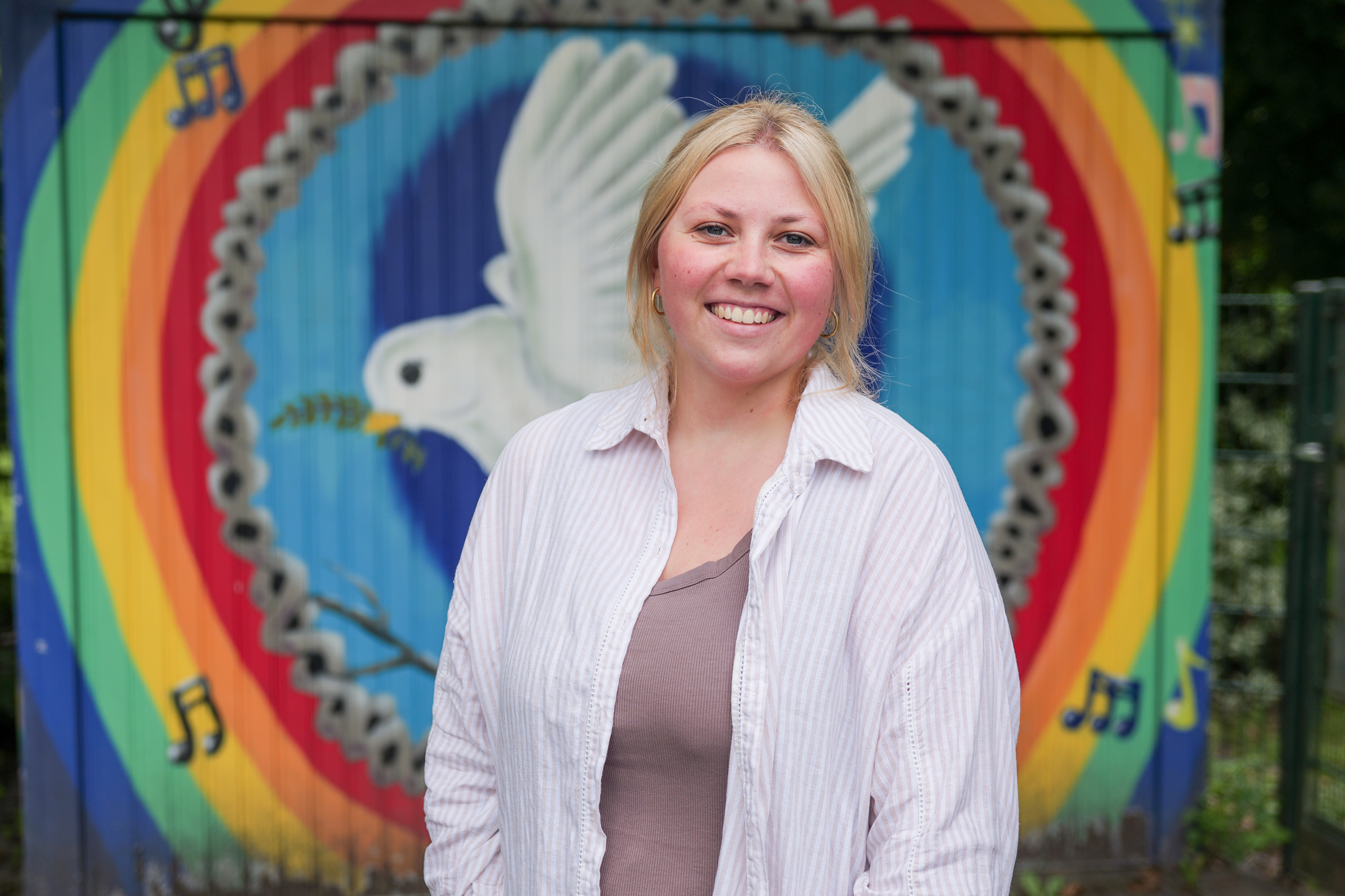 Eine junge Frau mit blonden Haaren und in einem weißen Hemd steht lächelnd vor einem bemalten Garagentor. Die Zeichnung zeigt eine Friedenstaube inmitten von Regenbogenfarben.