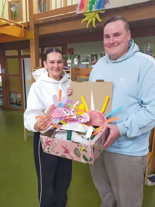 Zwei Personen stehen in einem Innenraum und halten eine geöffnete Box mit bunten, selbstgebastelten Papierarbeiten und Karten; im Hintergrund sind Fotos und Dekorationen zu sehen.