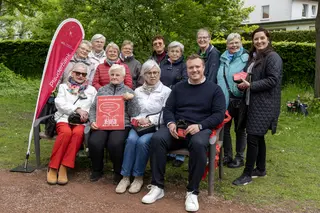 Gruppe von älteren und jüngeren Personen sitzt und steht um eine Bank in einem grünen Park; eine rote Fahne mit der Aufschrift „Plauderbank“ sowie ein Schild „Die CKD-Plauderbank – Wer hier sitzt, unterhält sich gerne!“ sind sichtbar.