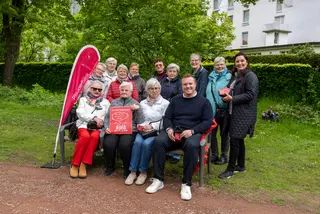 Gruppe von älteren und jüngeren Personen sitzt und steht um eine Bank in einem grünen Park; eine rote Fahne mit der Aufschrift „Plauderbank“ sowie ein Schild „Die CKD-Plauderbank – Wer hier sitzt, unterhält sich gerne!“ sind sichtbar.