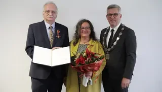 Das Bild zeigt zwei Männer und eine Frau. Der ältere Herr hält eine Urkunde ins Foto. Die Frau, Doratea Erkeling, hält einen Blumenstrauß.