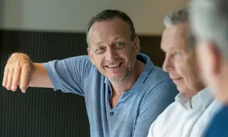 Mann schaut optimistisch in die Kamera. In der Unschärfe sind noch zwei Herren zu sehen.