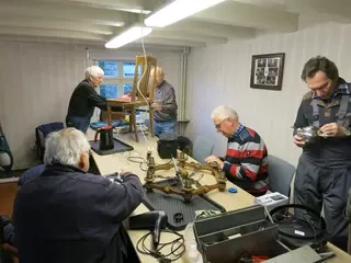 Fünf ältere Männer reparieren an einem Tisch verschiedene Gegenstände.