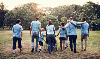 Das Bild zeigt eine Gruppe Menschen von hinten. Alle tragen hellblaue T-Shirts. Ein paar gehen Arm in Arm. In der Mitte sieht man, dass eine Person im Rollstuhl geschoben wird.