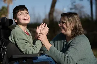 Ein Junge im Rollstuhl lacht. Seine Therapeutin umfasst seine Hände.