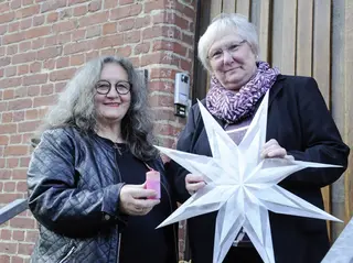 Zwei Frauen stehen auf einer Kirchentreppe. Eine Frau hält eine rosa Kerze ins Bild, die andere einen selbstgebastelten Stern.