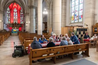 Das Bild zeigt eine Gruppe Menschen in einer Kirche, die zusammen beim Essen sitzen.