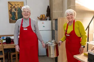 Das Bild zeigt zwei Frauen mit roten Schürzen, die einen Suppentopf halten.