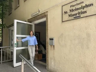 Das Bild zeigt eine bebrillte Frau, die die Tür zu einem Pfarrheim offen hält.