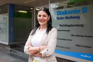 Eine Frau steht vor einem Büro der Diakonie. Sie hat schwarze Haare und trägt einen beigen Blazer über einem weißen Hemd.